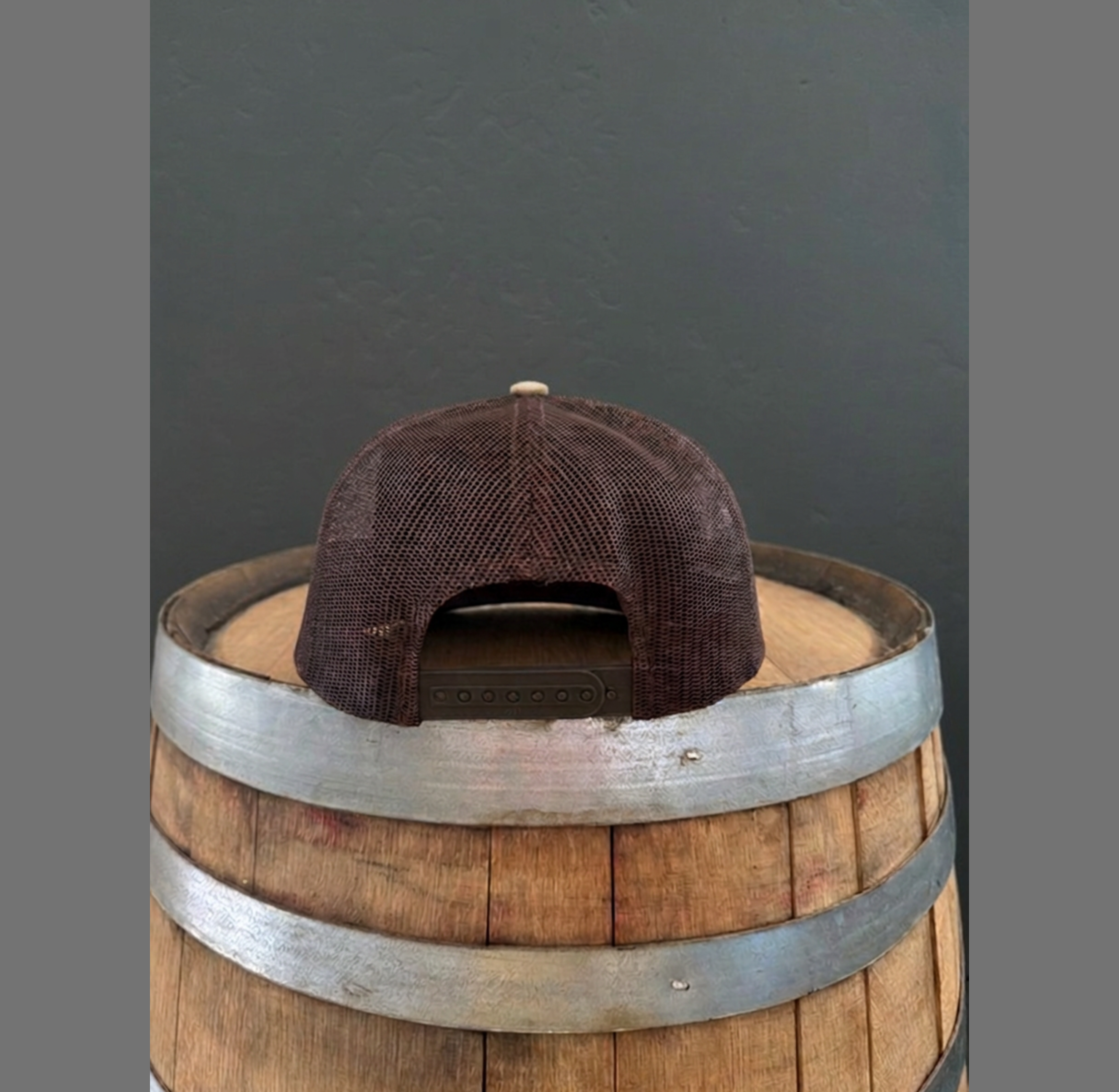 Back view of Mustache Ranch Coffee Co. camo brown trucker hat showing dark brown snapback closure and mesh back on a wooden barrel