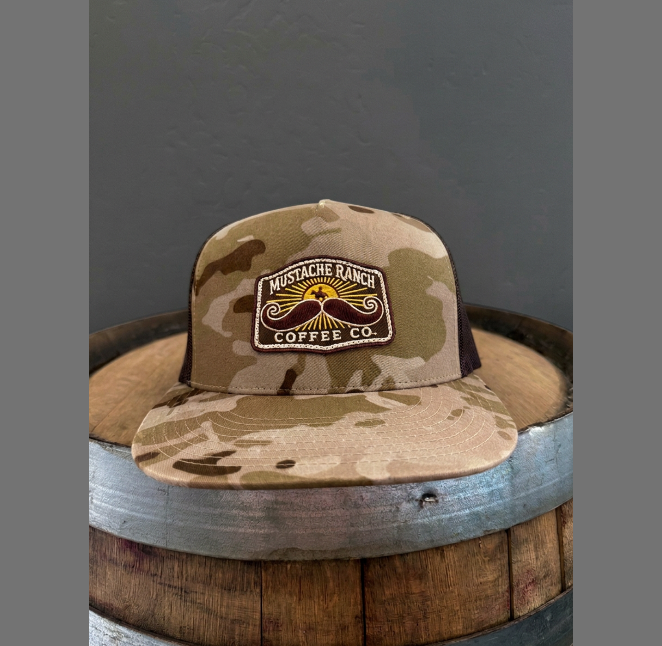 Mustache Ranch Coffee Co. camo brown flat bill trucker snapback hat with front patch on a wooden barrel