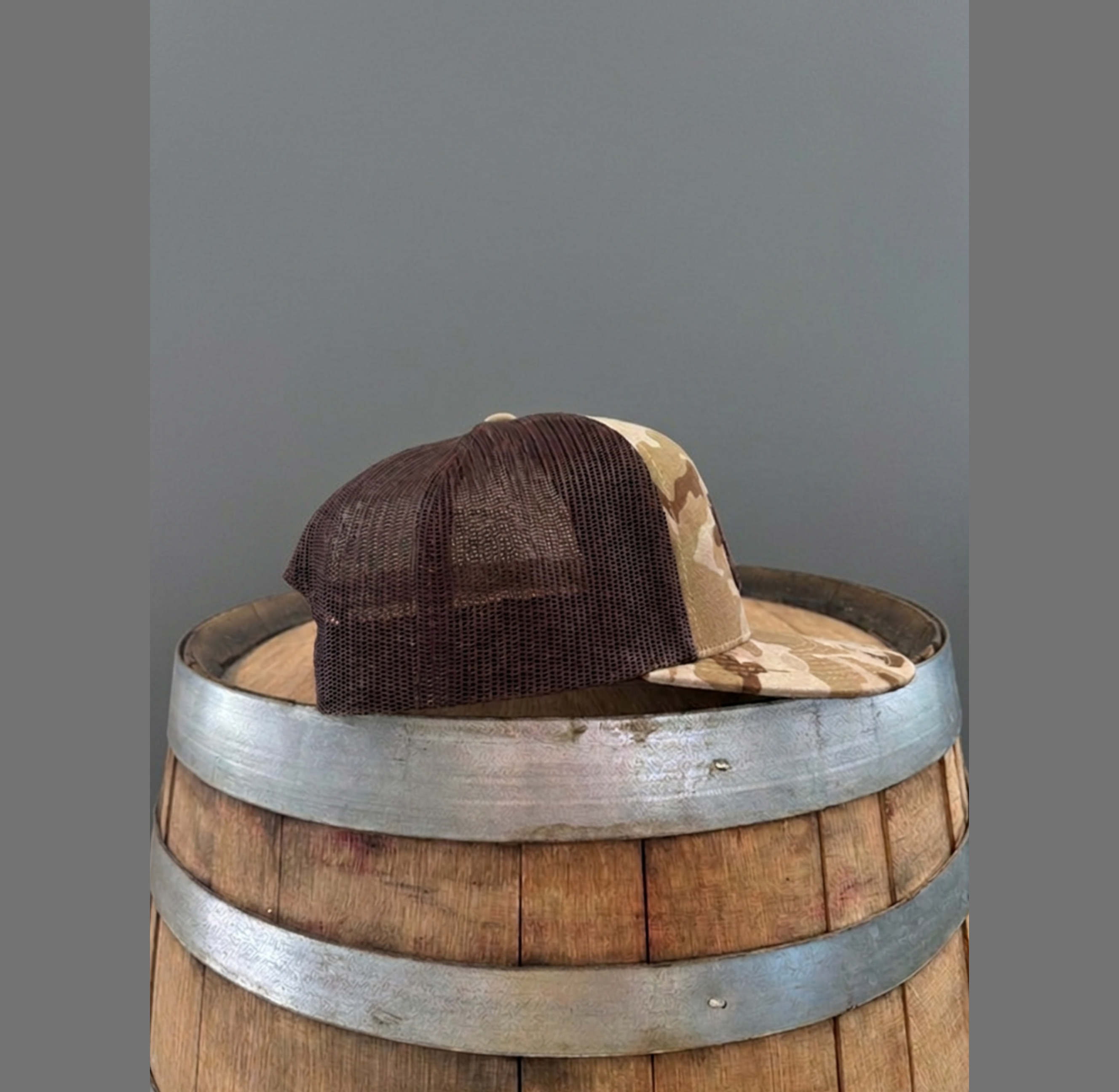 Side profile view of Mustache Ranch Coffee Co. camo brown trucker snapback hat showing dark brown mesh netting and flat bill on a wooden barrel