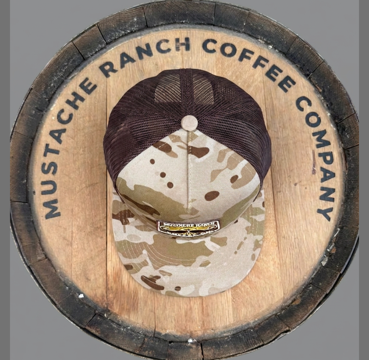 Top-down view of Mustache Ranch Coffee Co. camo brown trucker snapback hat displayed on a branded wooden barrel lid