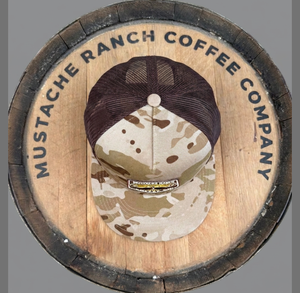 Top-down view of Mustache Ranch Coffee Co. camo brown trucker snapback hat displayed on a branded wooden barrel lid