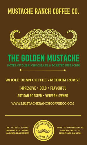 A rich brown coffee label with an intricate gold filigree handlebar mustache. The text "The Golden Mustache" is highlighted in green with subtext for Dubai chocolate and toasted pistachio flavor notes.