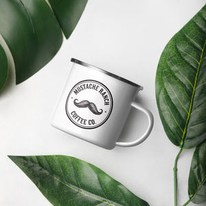 Top-down view of the 12 oz. Mustache Ranch Coffee Co. white enamel mug with a silver rim, featuring the black logo, styled on a white background with decorative green tropical leaves.