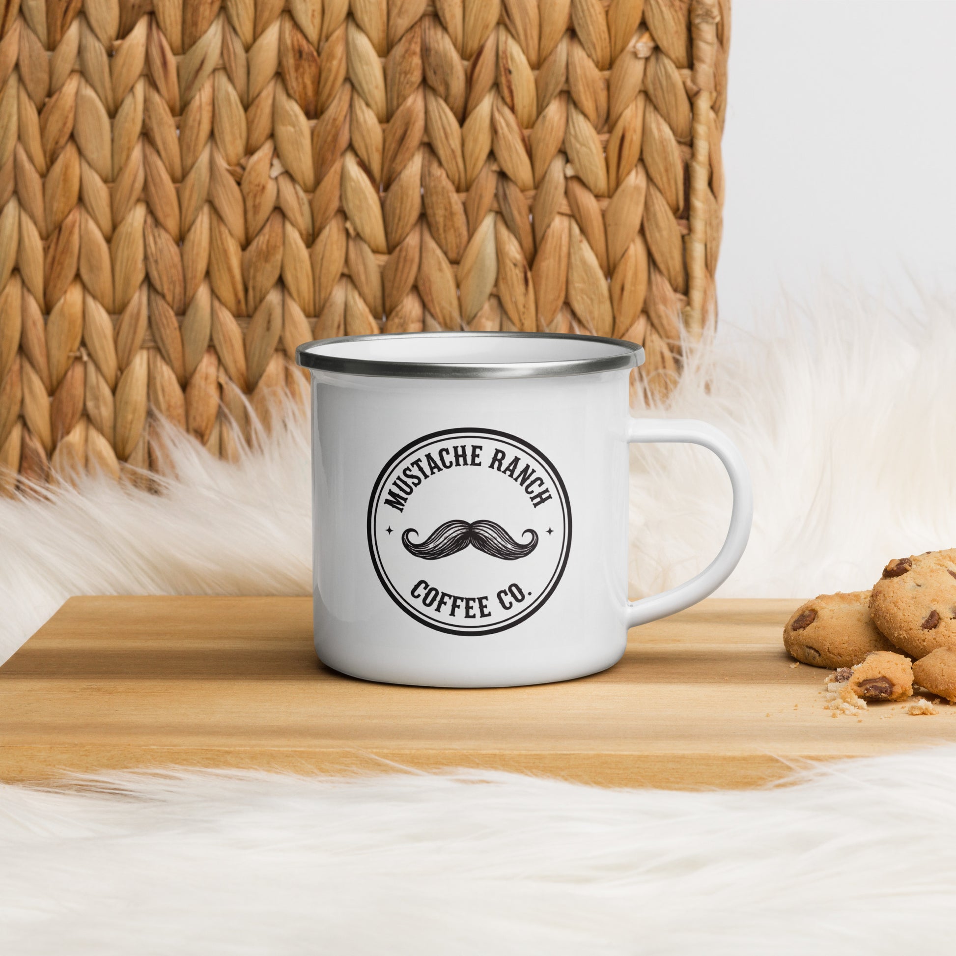 White 12 oz. Mustache Ranch Coffee Co. enamel mug with black logo, displayed on a wooden surface with a woven basket and cookies, styled for a rustic, farmhouse aesthetic.